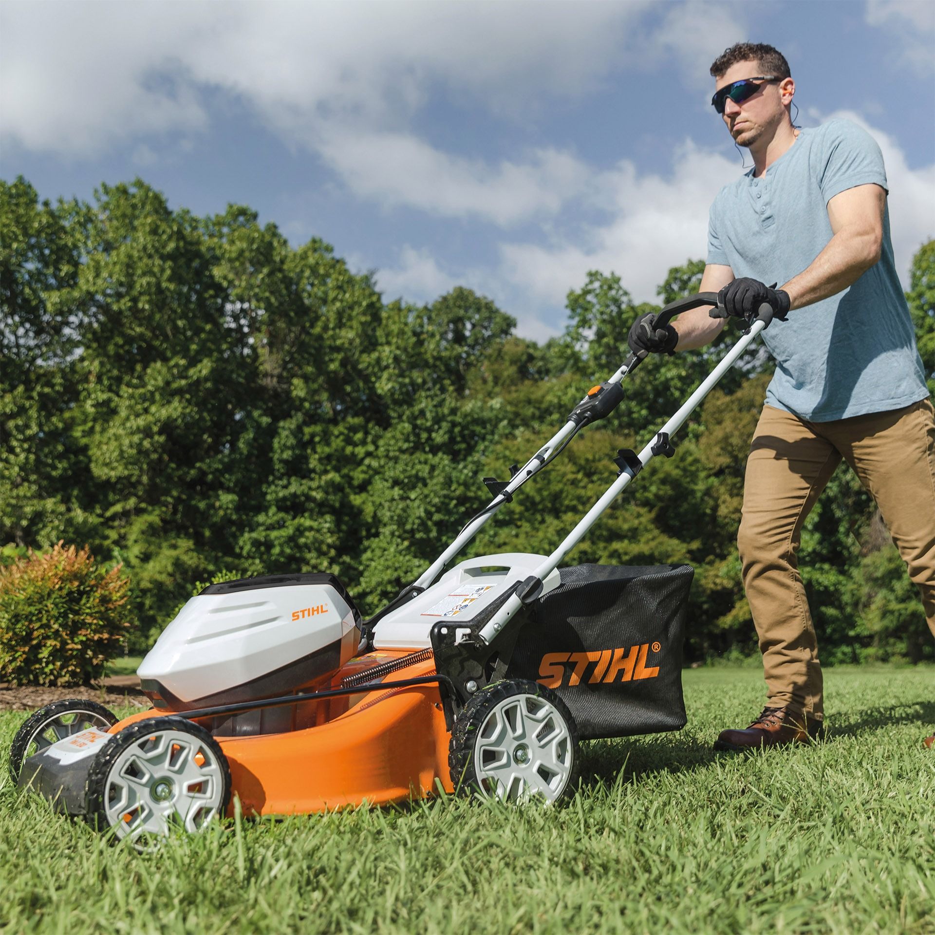 Man mowing grass with an orange and white lawnmower, in a sunny yard with trees.