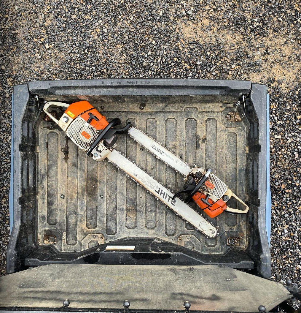 Two chainsaws in a truck bed, against a dirt backdrop. Orange and white.