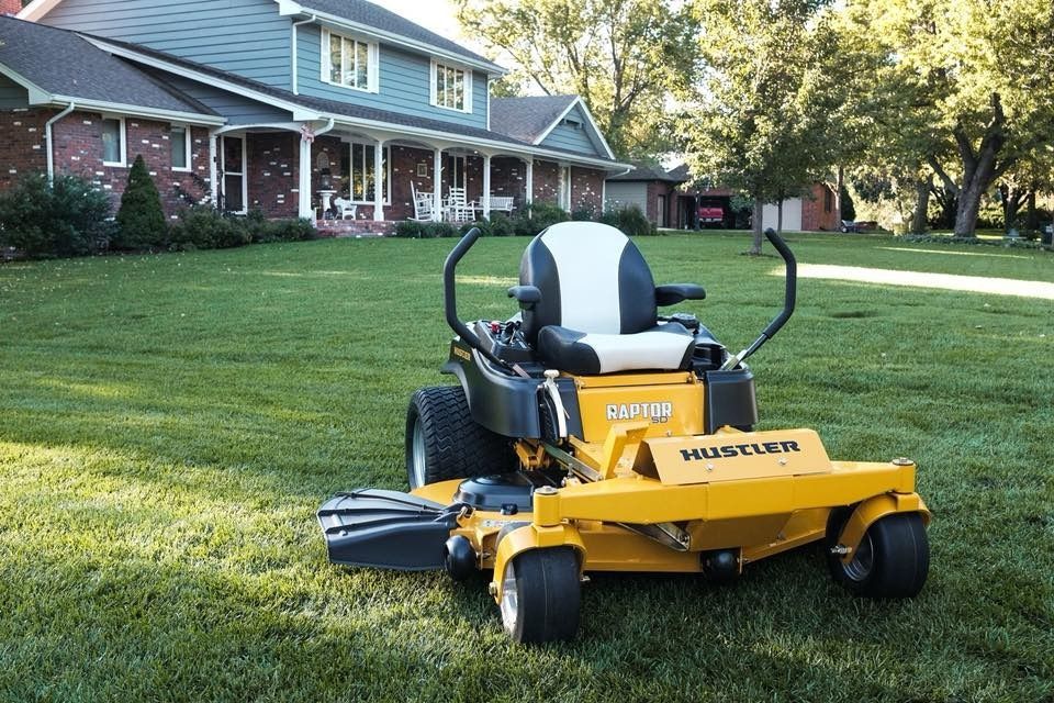 Yellow Hustler zero-turn mower on a green lawn in front of a house.