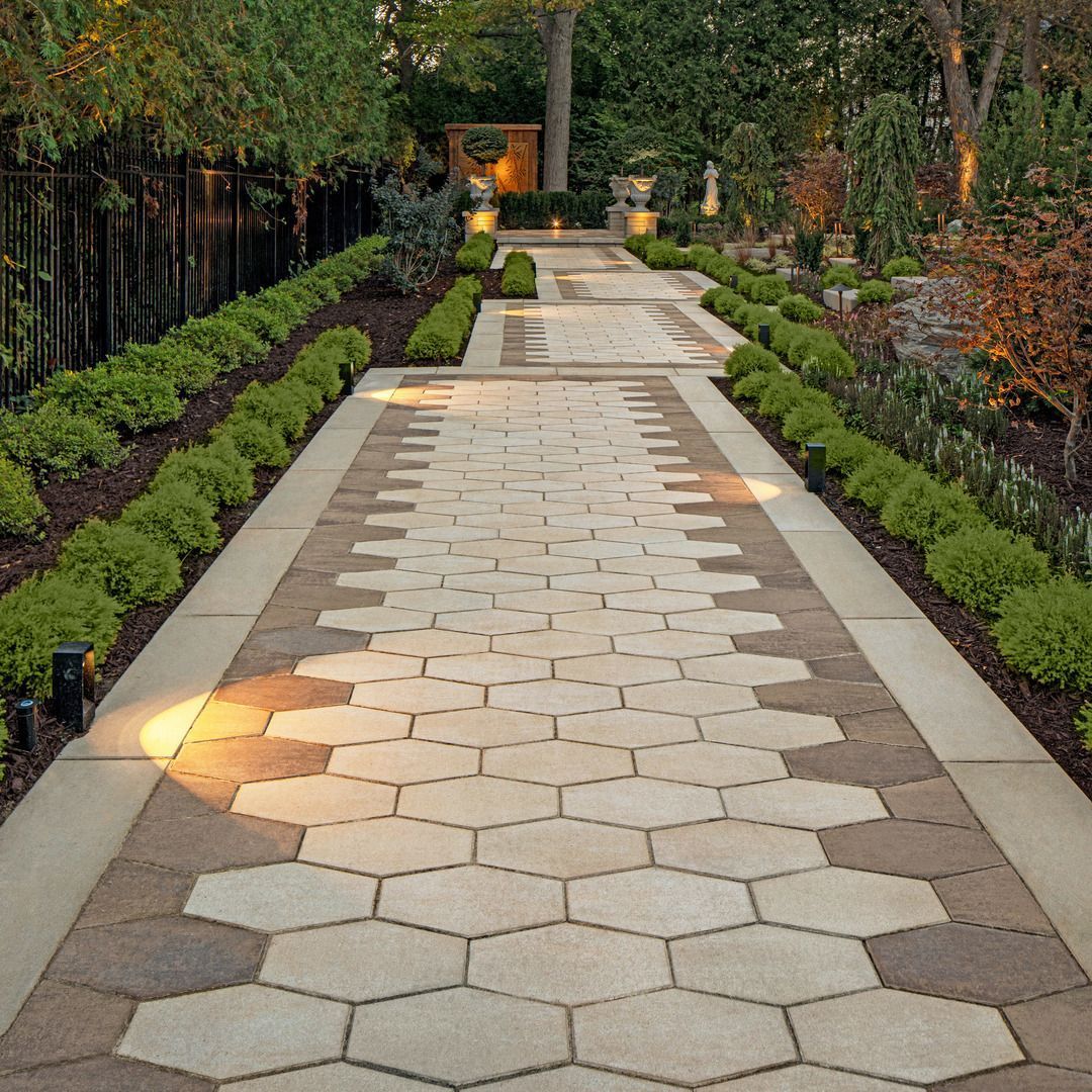 Stone garden path bordered by hedges and flowers, leading to a lit doorway in a landscaped yard