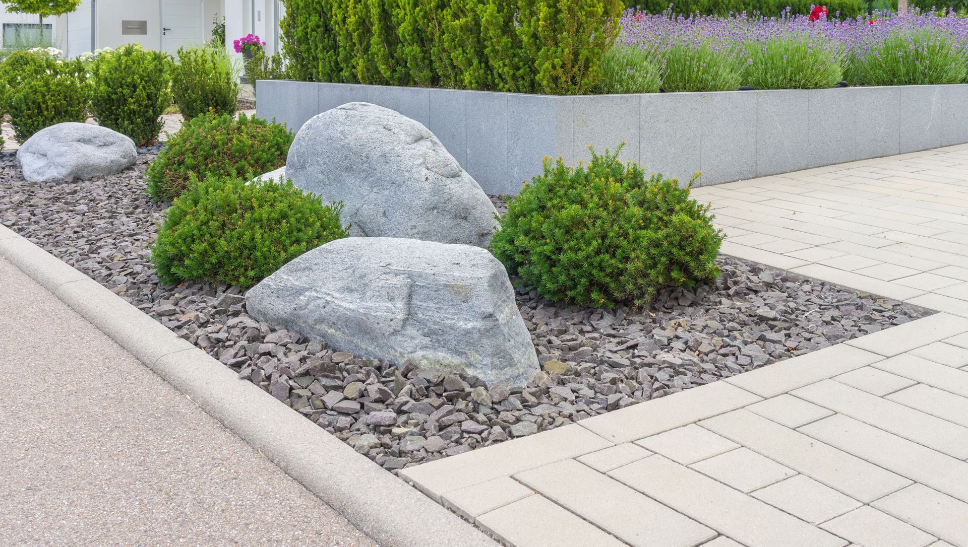 Landscaped garden bed with large rocks, shrubs, and gravel beside a paved walkway