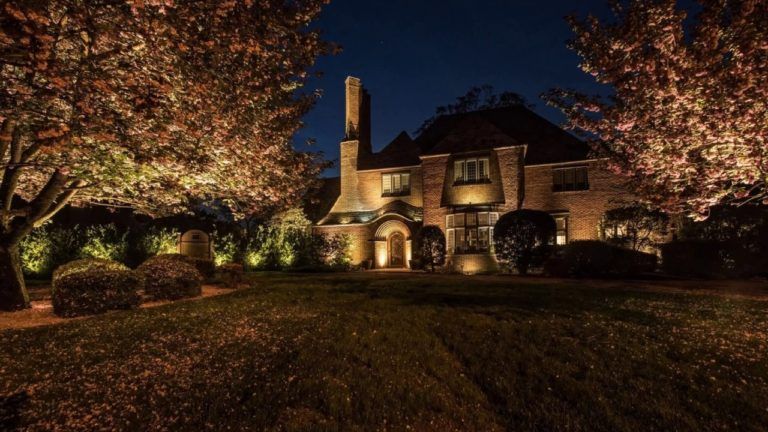 Illuminated stone house at night, framed by lit trees and a dark lawn