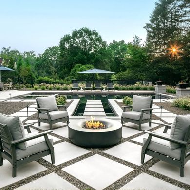 Modern patio with gray lounge chairs around a round fire pit, bordered by greenery at sunset