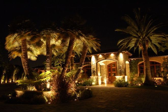 Illuminated stone house at night, framed by lit trees and a dark lawn