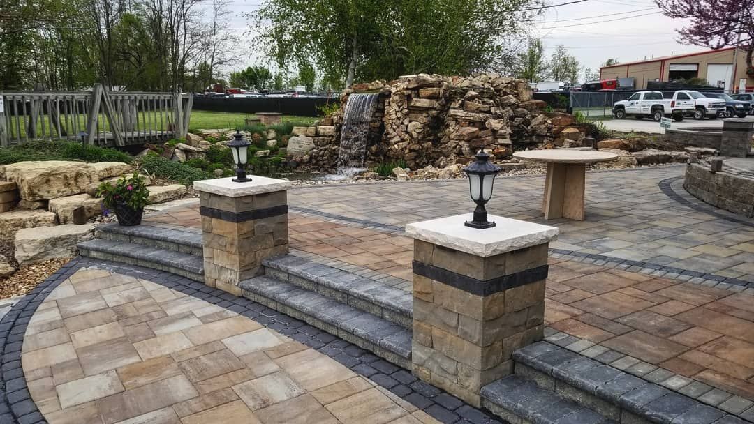 Stone steps and brick patio with two decorative lamps, leading to a small rock waterfall in a landscaped plaza.