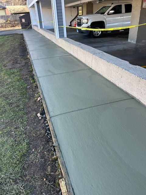 Freshly poured concrete sidewalk next to a building, yellow caution tape, and a white pickup truck.