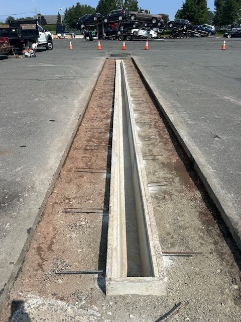 Concrete drainage channel in asphalt parking lot, with metal bars and surrounding dirt, cars in background.