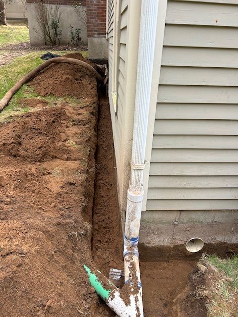 Trench dug alongside a house, next to a white gutter pipe. Dirt pile and grass visible.
