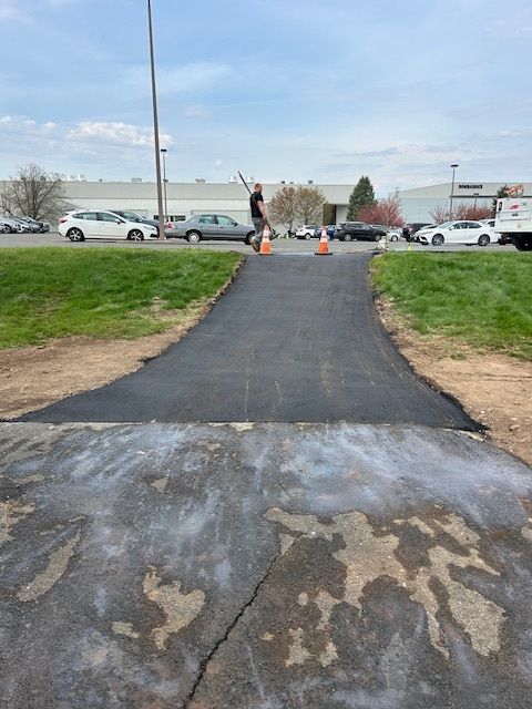 New asphalt ramp leading to a parking lot. A person gestures near the ramp. Cars and a building are in the background.