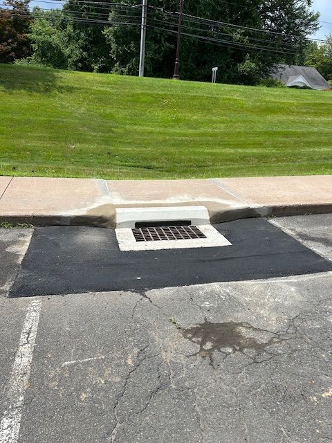Drain in asphalt parking lot next to a curb and grassy area. The surrounding asphalt is cracked.