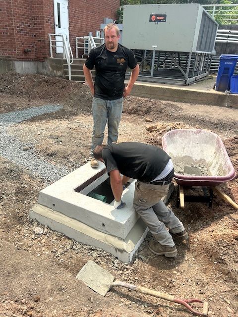 Two construction workers installing a concrete access box outdoors. One stands, the other leans into the opening. A shovel and wheelbarrow are nearby.