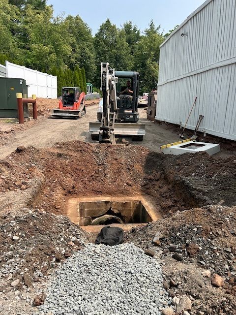 Construction site with an excavator digging into a square hole near a building.