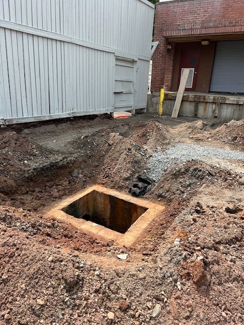Construction site with an open, concrete-lined pit. Dirt and debris surround the pit near a building.