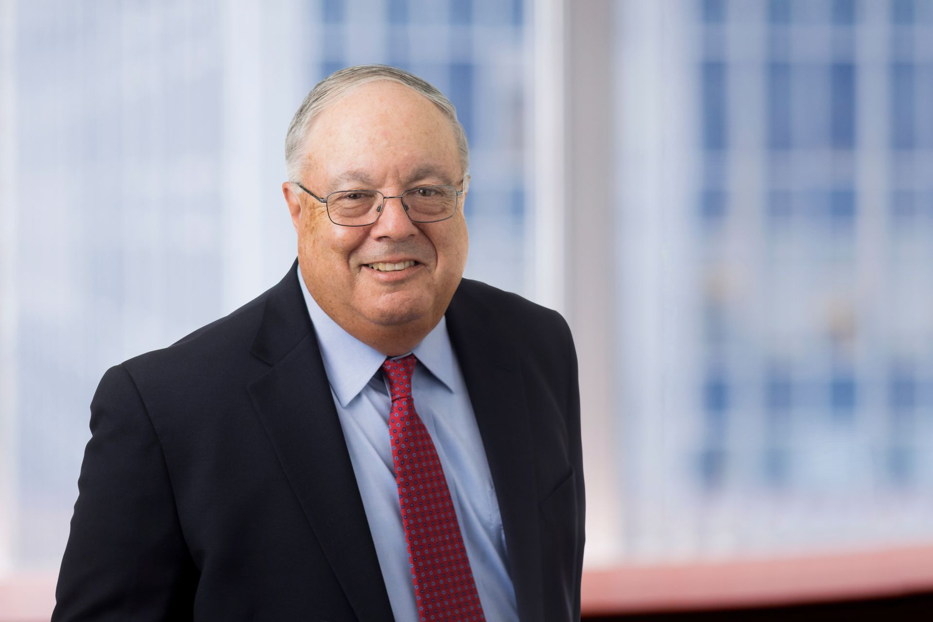 Man in suit smiles, wearing glasses and a red tie, in a business setting with a window in the background.