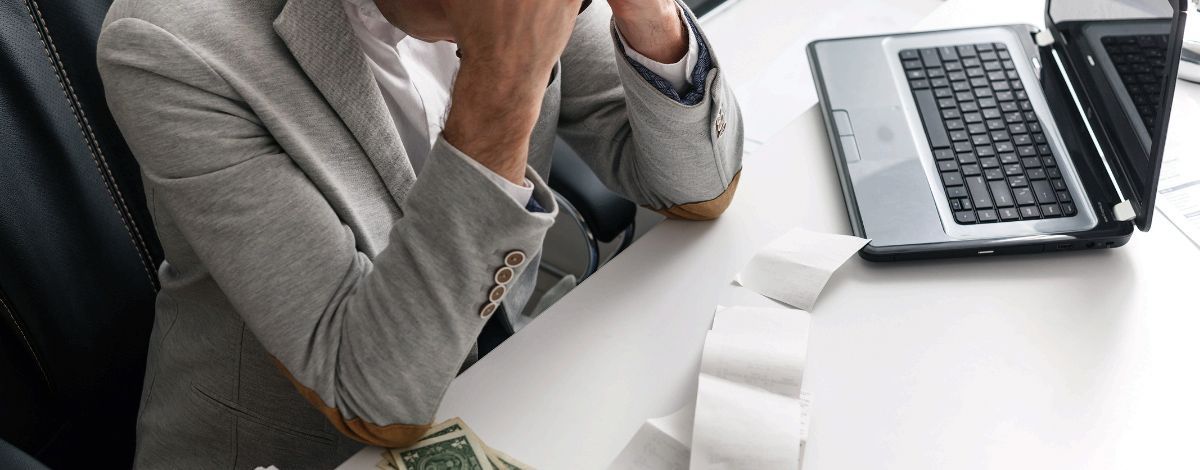 Man in grey blazer stressed at desk with laptop, receipts, and money.