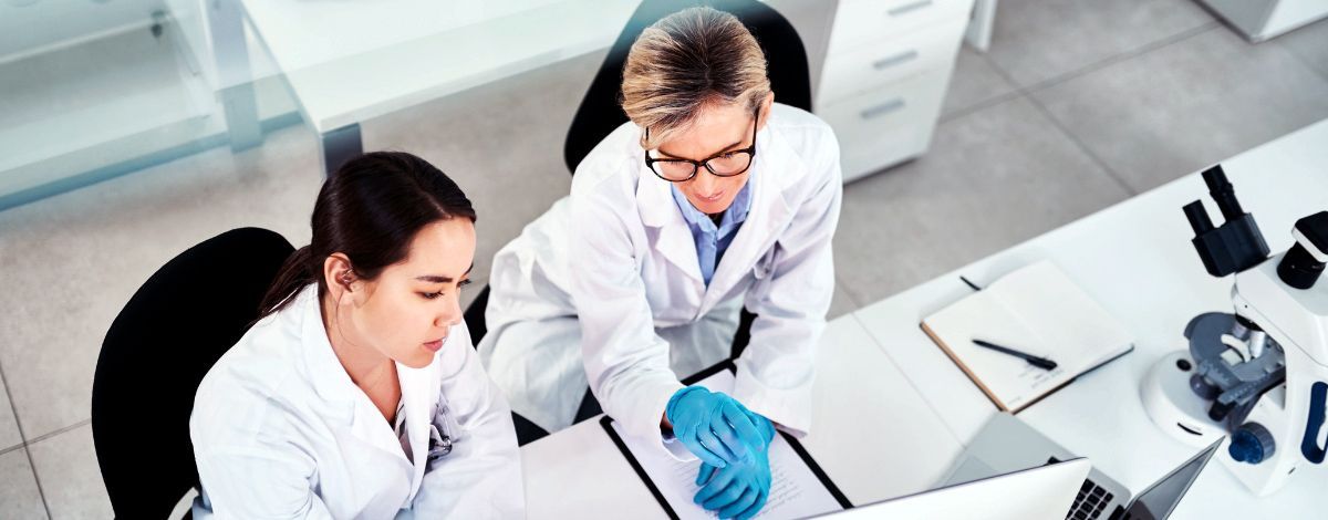 Two scientists in lab coats examine data on a computer in a lab setting.