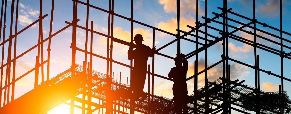 Silhouetted construction workers on scaffolding against a sunset sky.