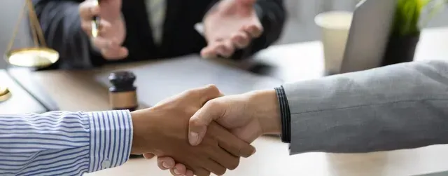 Two people shaking hands at a desk, possibly a legal setting, with a gavel and scales visible.