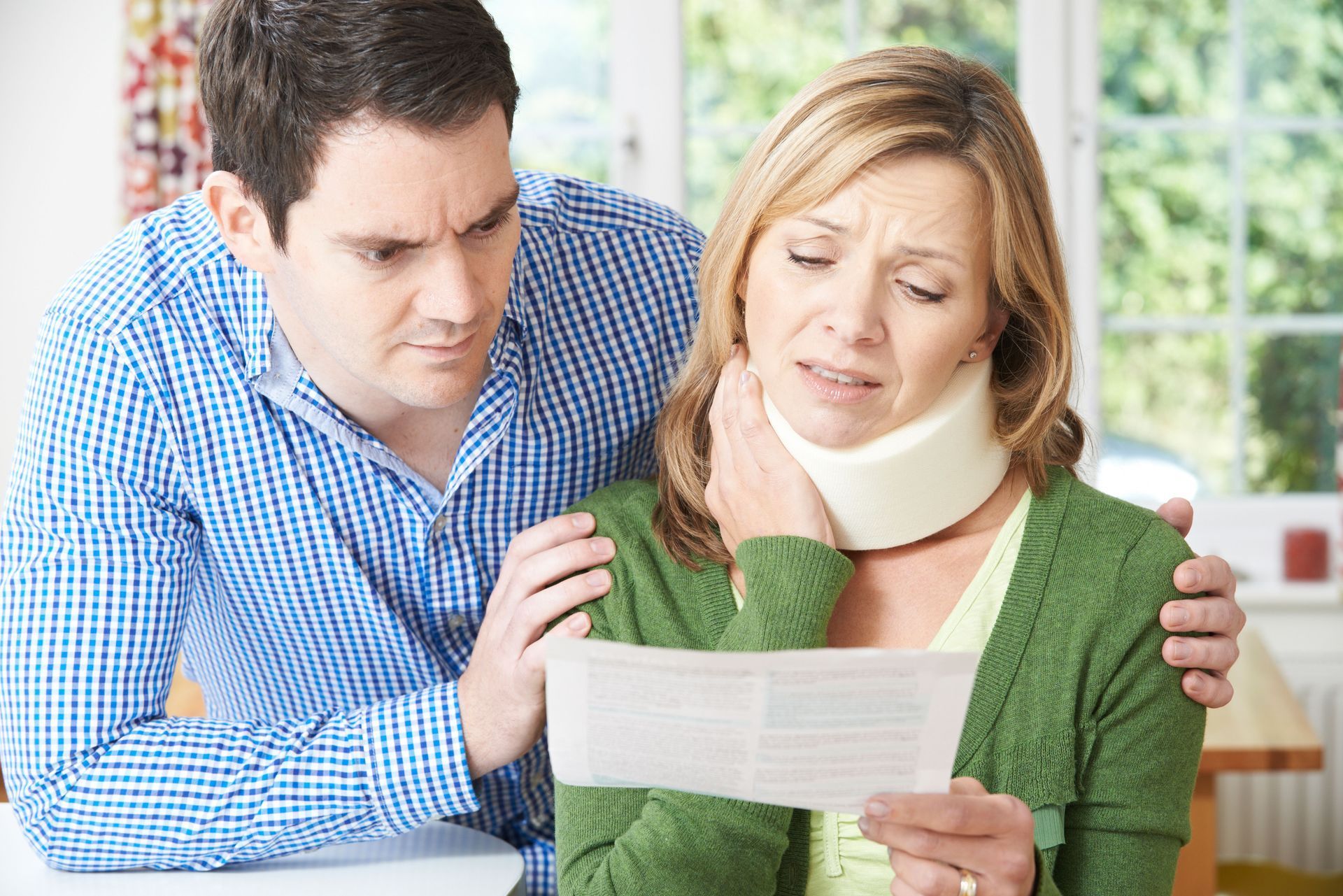 Woman in neck brace, looking distressed, reads a document; man comforts her.