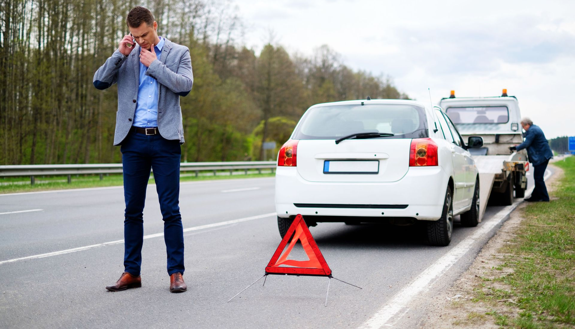 Man on phone by a white car being towed on the side of a highway. Red triangle safety sign present.