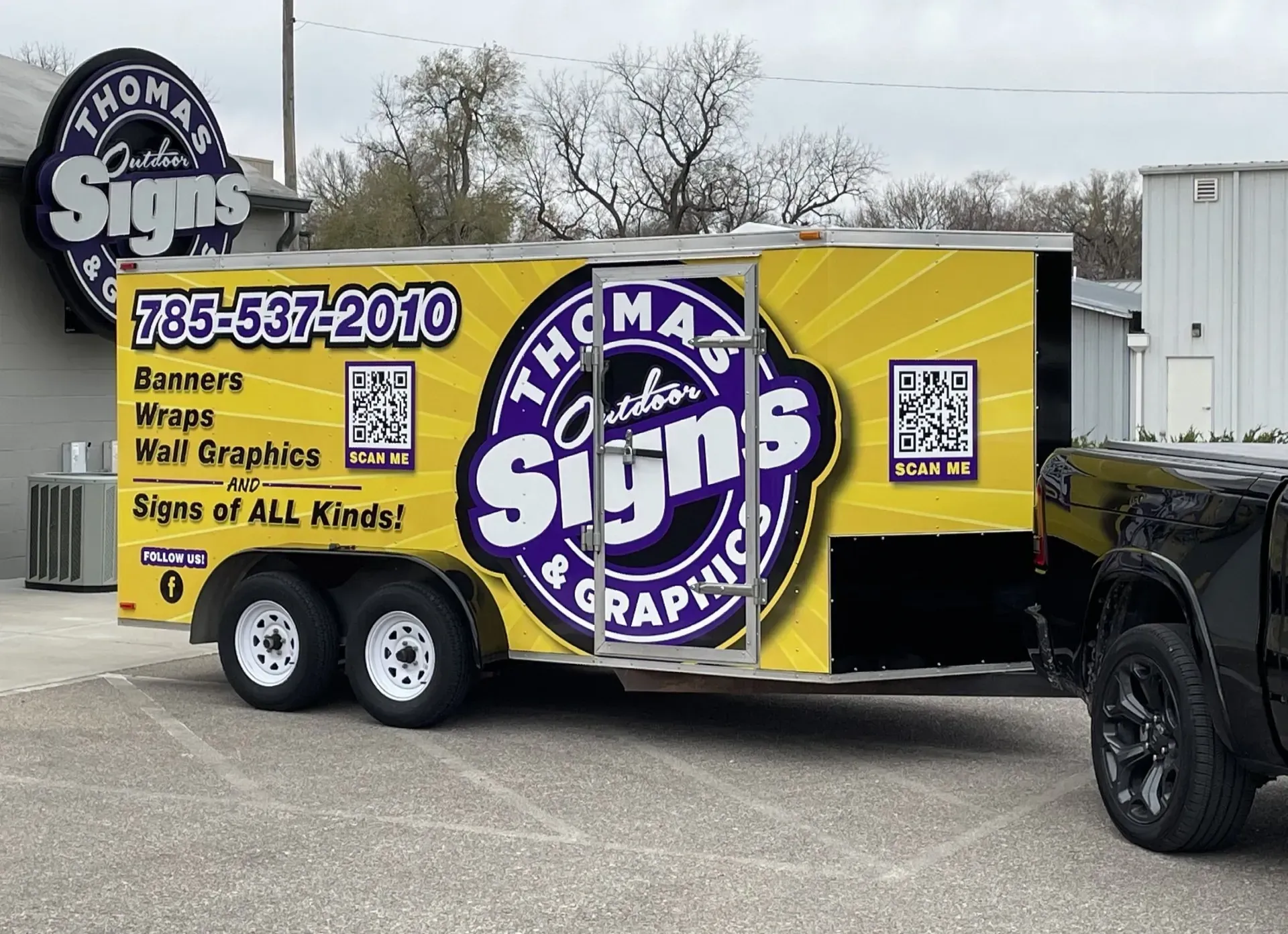 A yellow trailer with "Thomas Custom Signs & Graphics" logo. It's parked next to a building and a black truck.