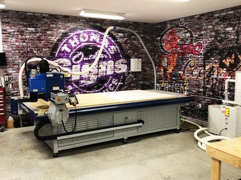 CNC router in a workshop with graffiti-covered brick wall and Thomas' Outfitters logo.