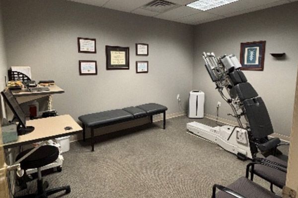 Medical exam room with a treatment table, adjustable chairs, and framed certificates on gray walls.