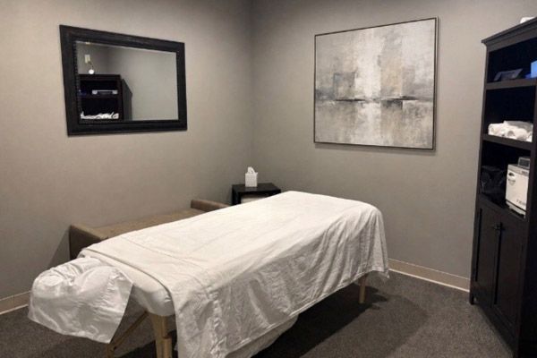 Massage table in a small treatment room with a mirror, wall art, and shelves.
