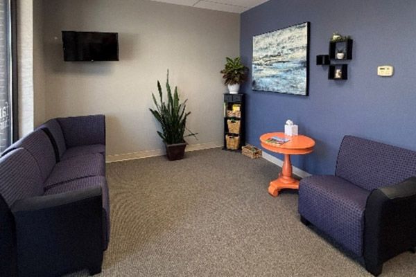 Small waiting room with purple chairs, wall art, a plant, and a round orange table.