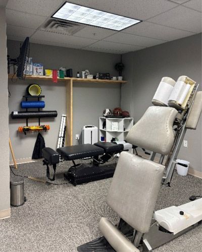 Physical therapy room with treatment table, exercise equipment, shelves, and a ceiling light panel.
