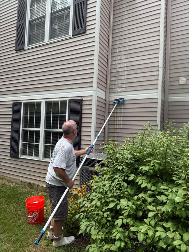 A person uses a long-handled brush to clean the grey vinyl siding of a two-story house near a shrub and red bucket.