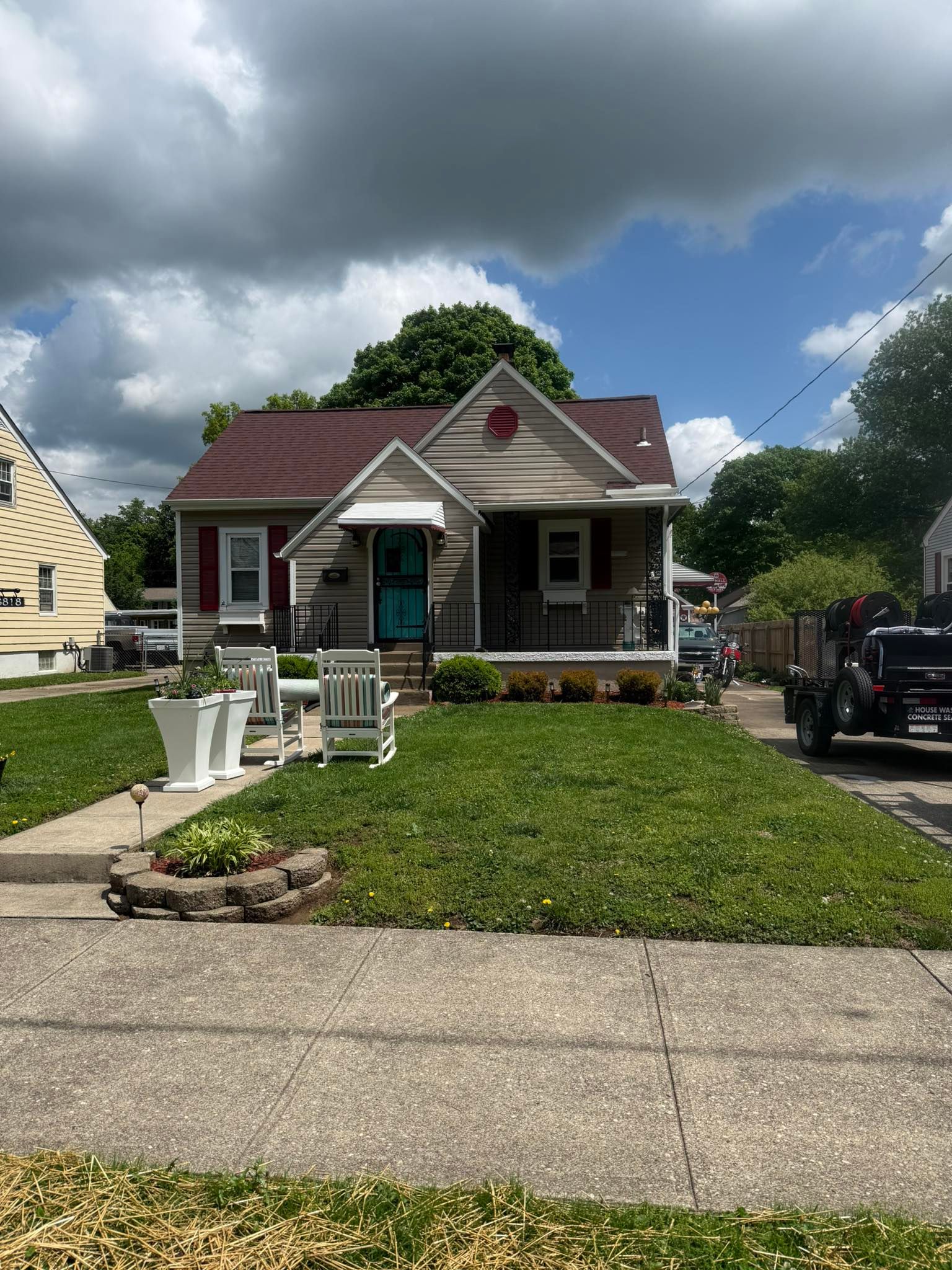 Small white house with red trim and porch, front lawn, cloudy sky, and parked vehicles on the street.