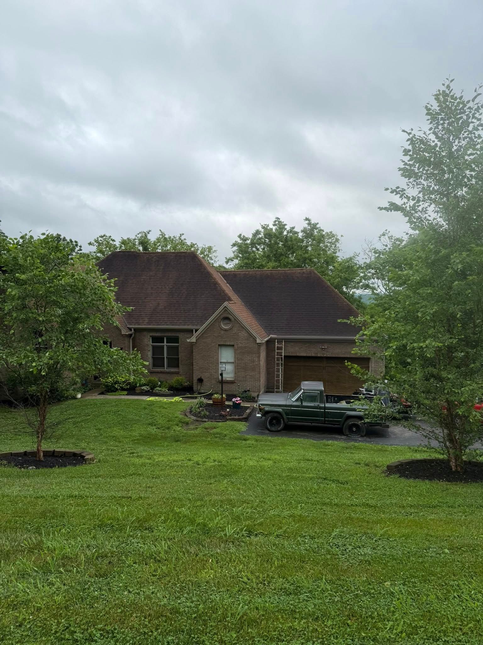 Suburban brick house with a dark car in the driveway, surrounded by green lawn and trees under cloudy skies
