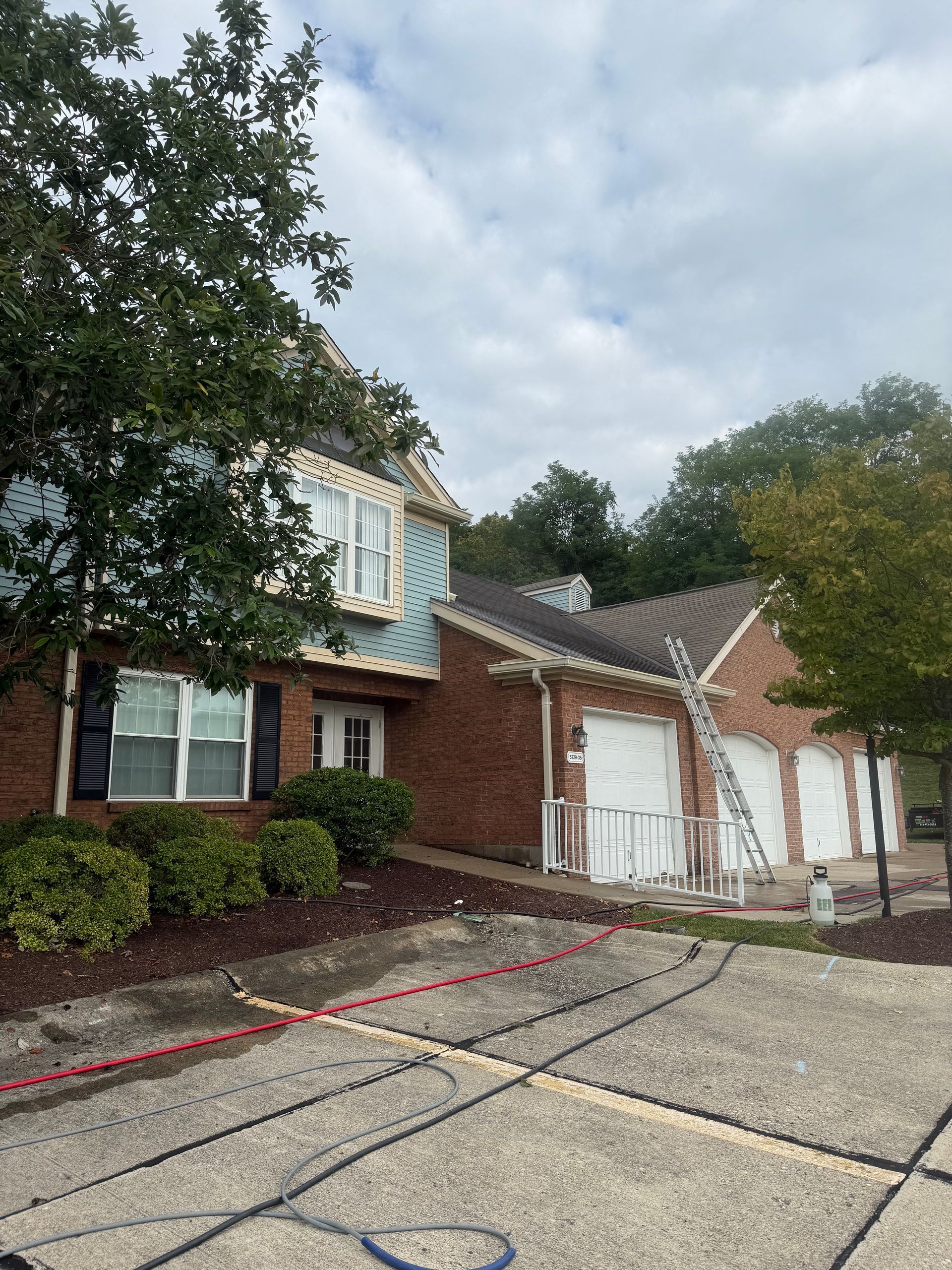 Brick suburban house with white garage doors, gravel driveway, and trees under a cloudy sky