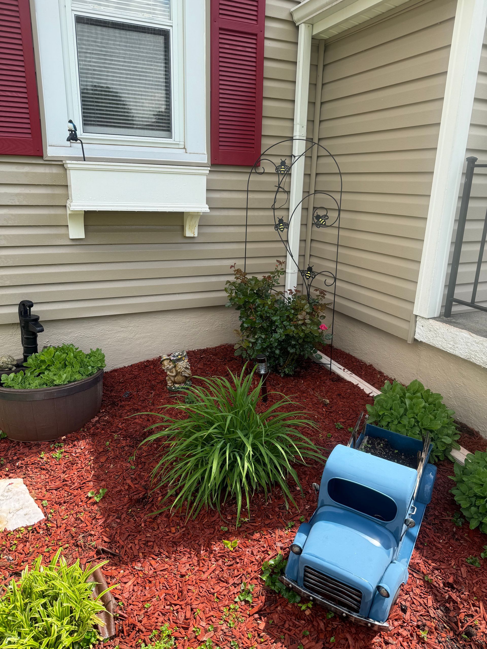 Small front garden with red mulch, green plants, a beige house wall, and a blue plastic step stool.