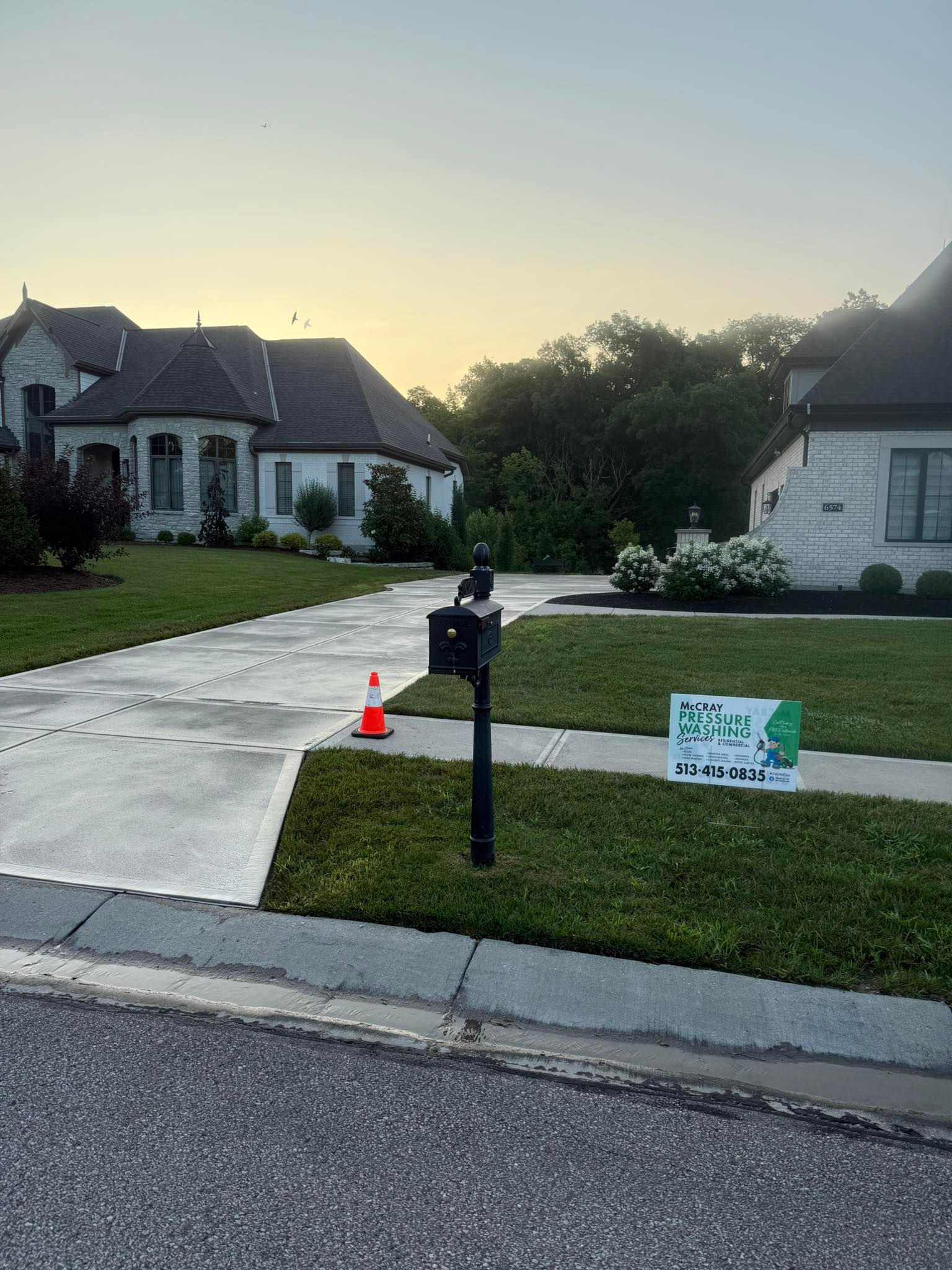 Suburban house at dusk with a driveway, mailbox, orange cone, and a lawn sign by the curb.