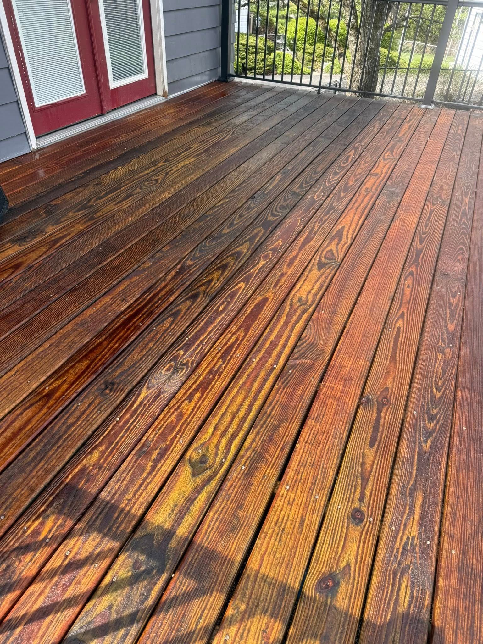 Weathered wooden porch deck with dark stains and sunlight shadows beside red doors and a black railing