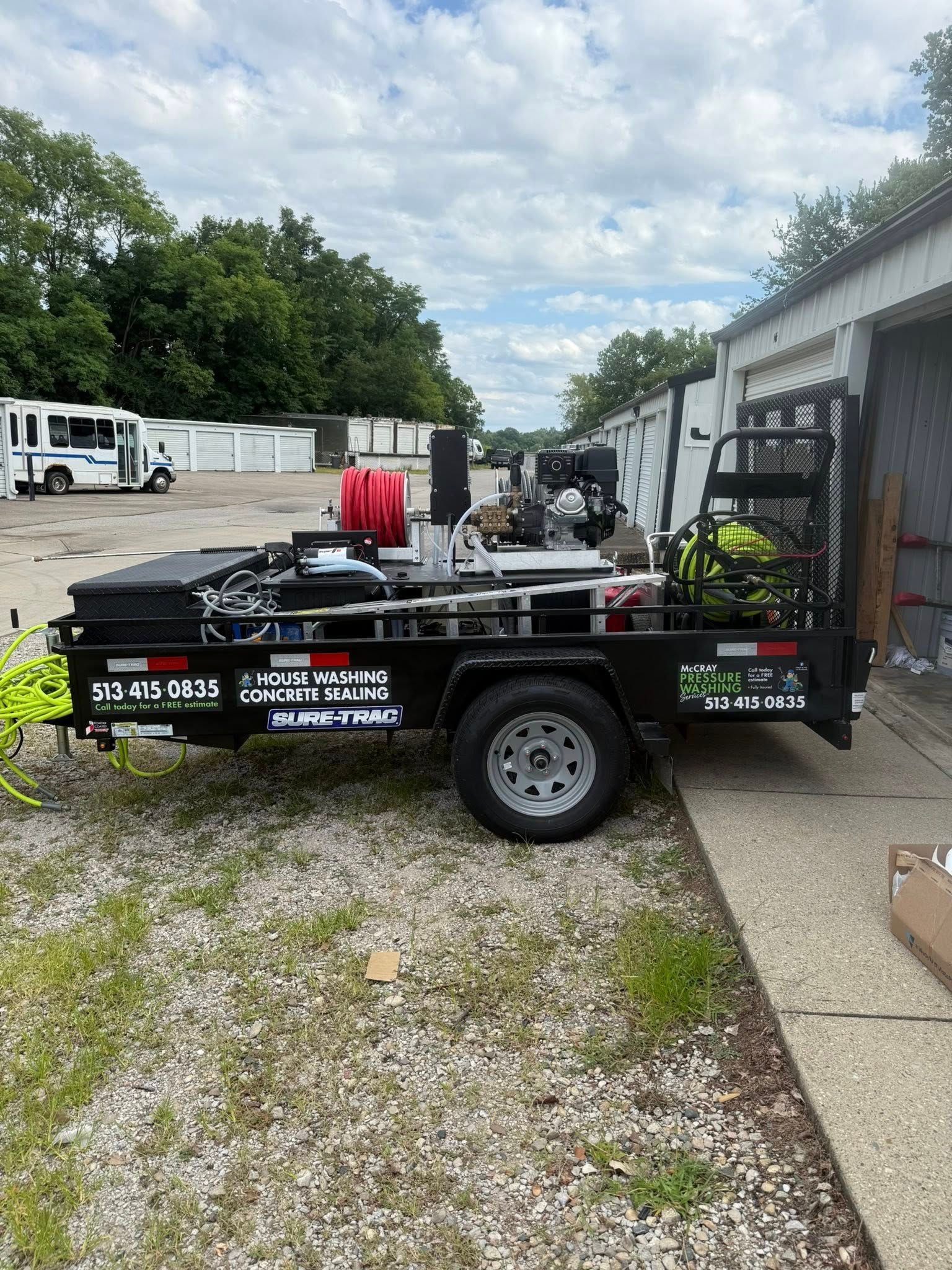 Black flatbed utility trailer with equipment parked beside a warehouse on a gravel lot