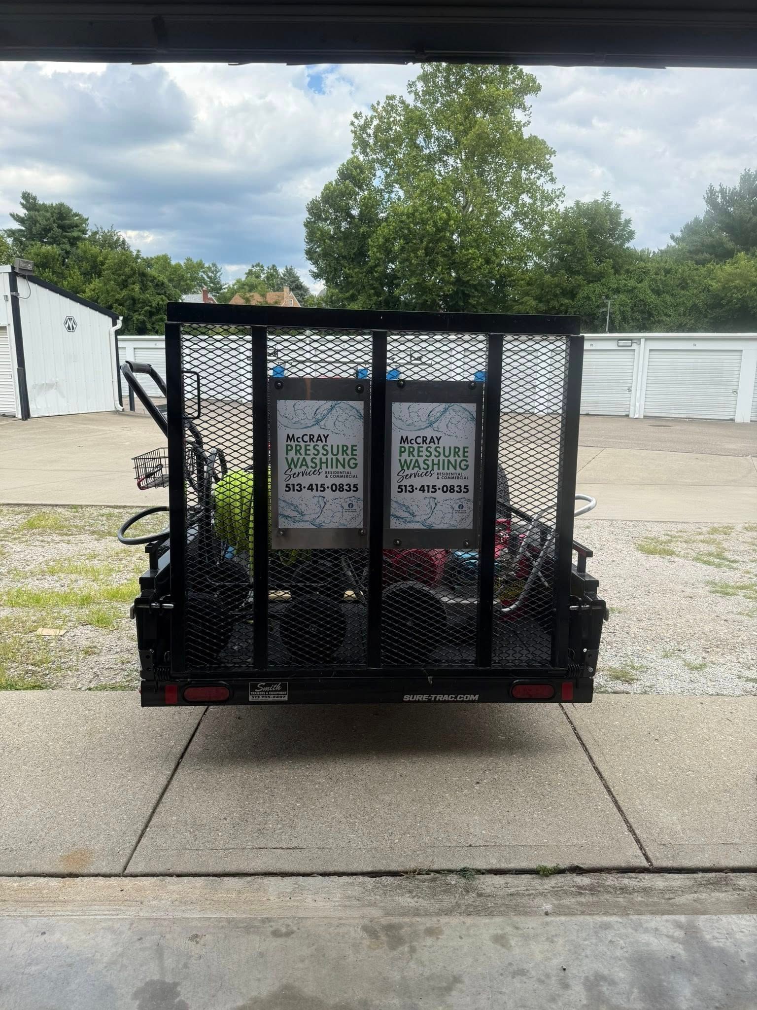 Black metal utility trailer on a concrete lot, with posters and tools inside, trees and white buildings behind.