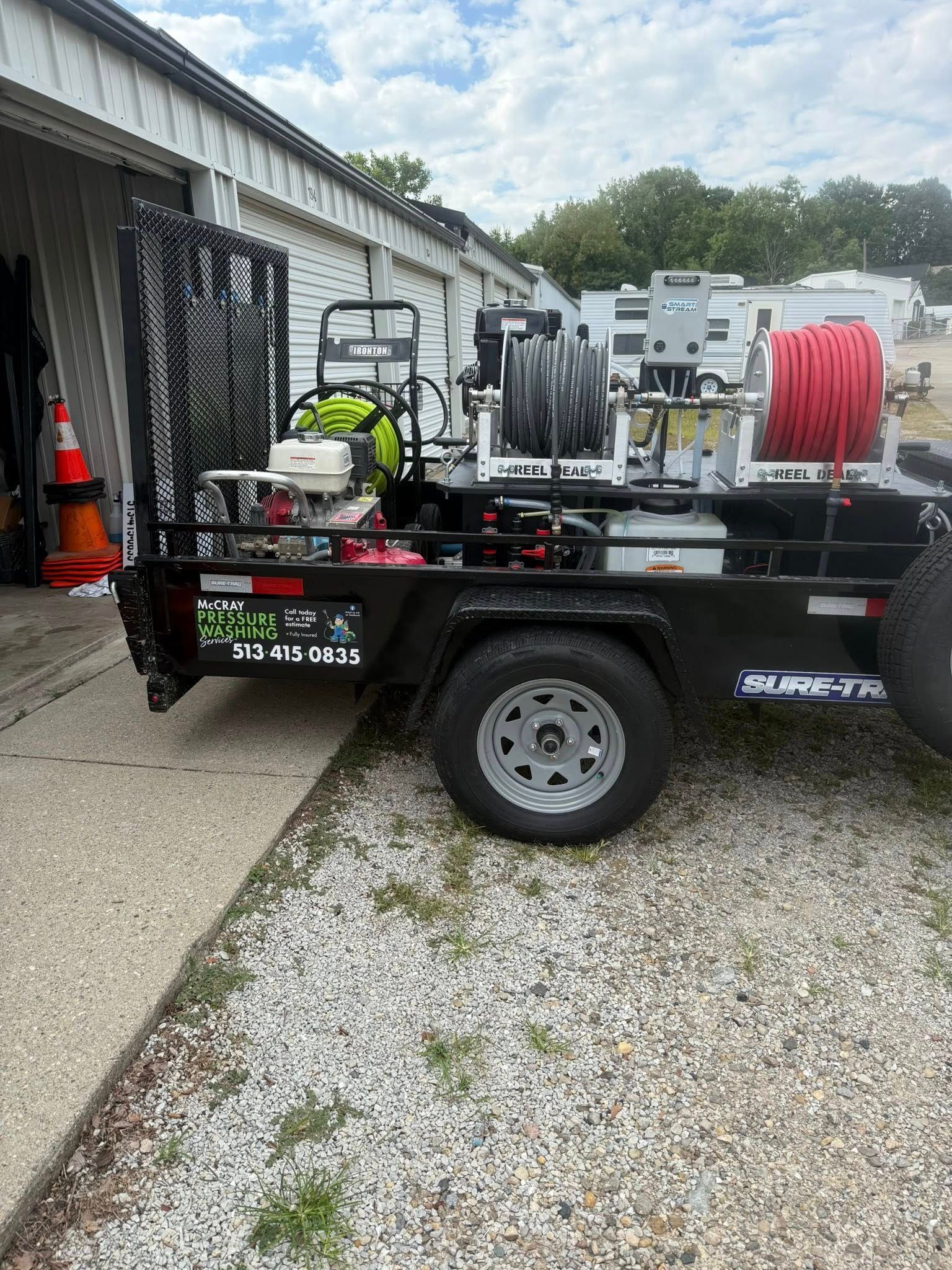 Utility trailer with a pressure washer, hoses, and equipment parked beside a building on gravel.