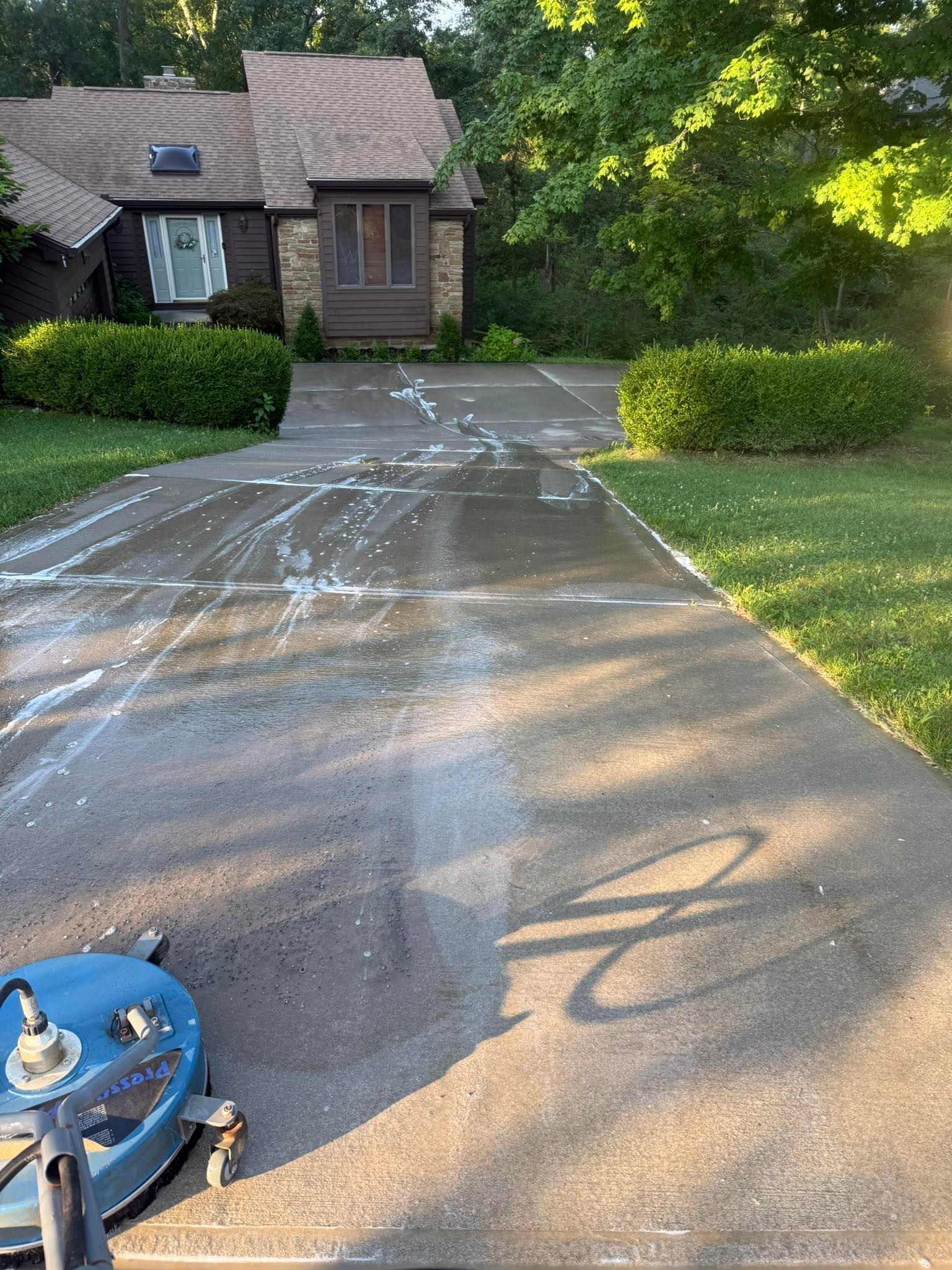 Driveway with wet streaks in front of a brick house, sunlight, and a blue pressure washer at the lower left