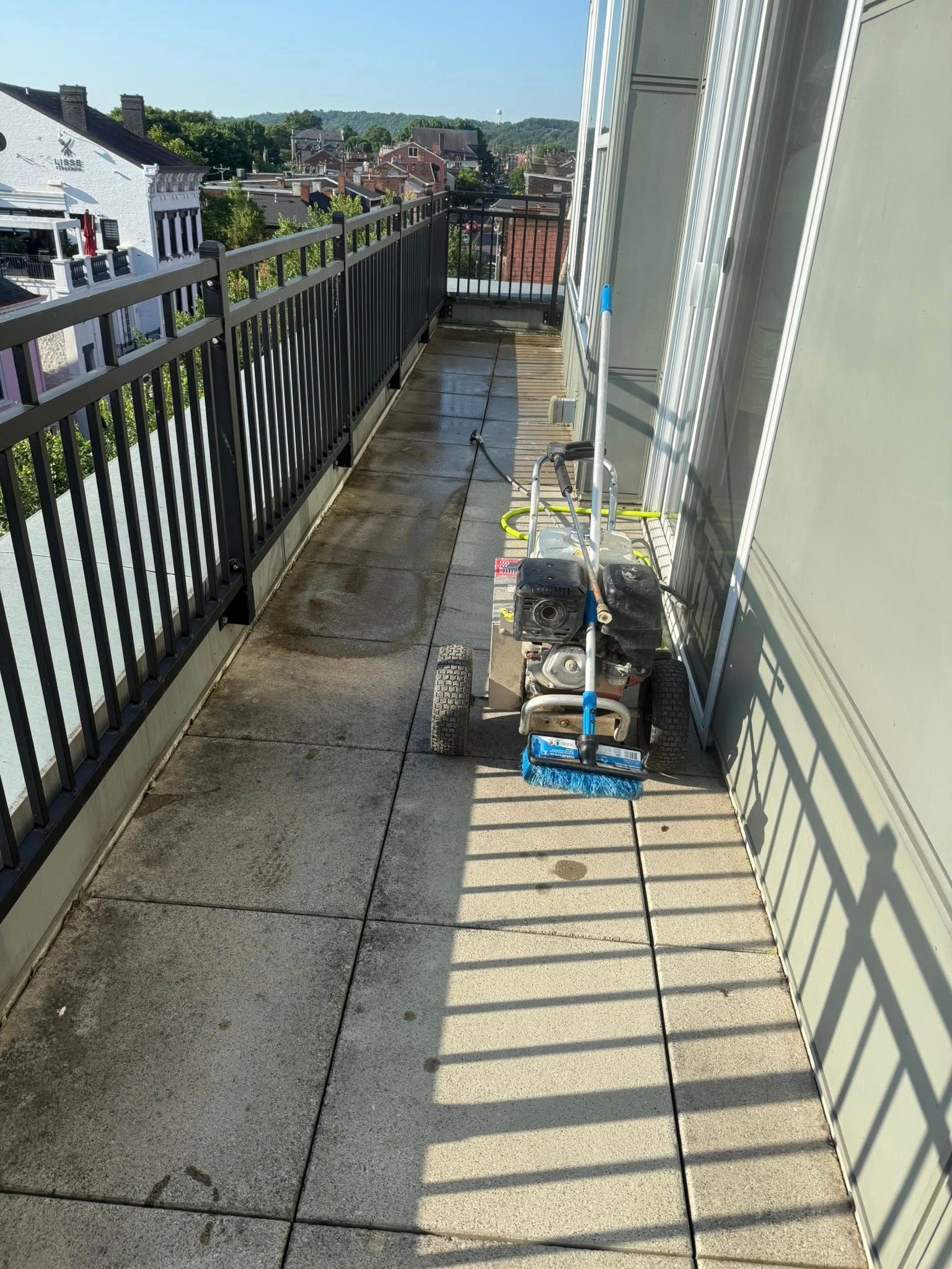 Narrow apartment balcony with black railing, cleaning supplies, and sunlight casting shadows.