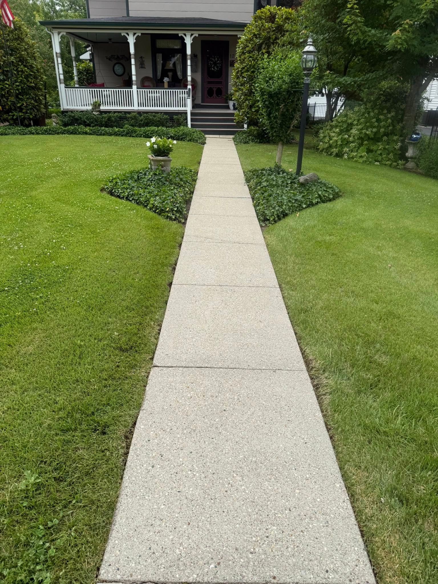 Front walkway leading to a porch on a house, bordered by green lawns and shrubs.