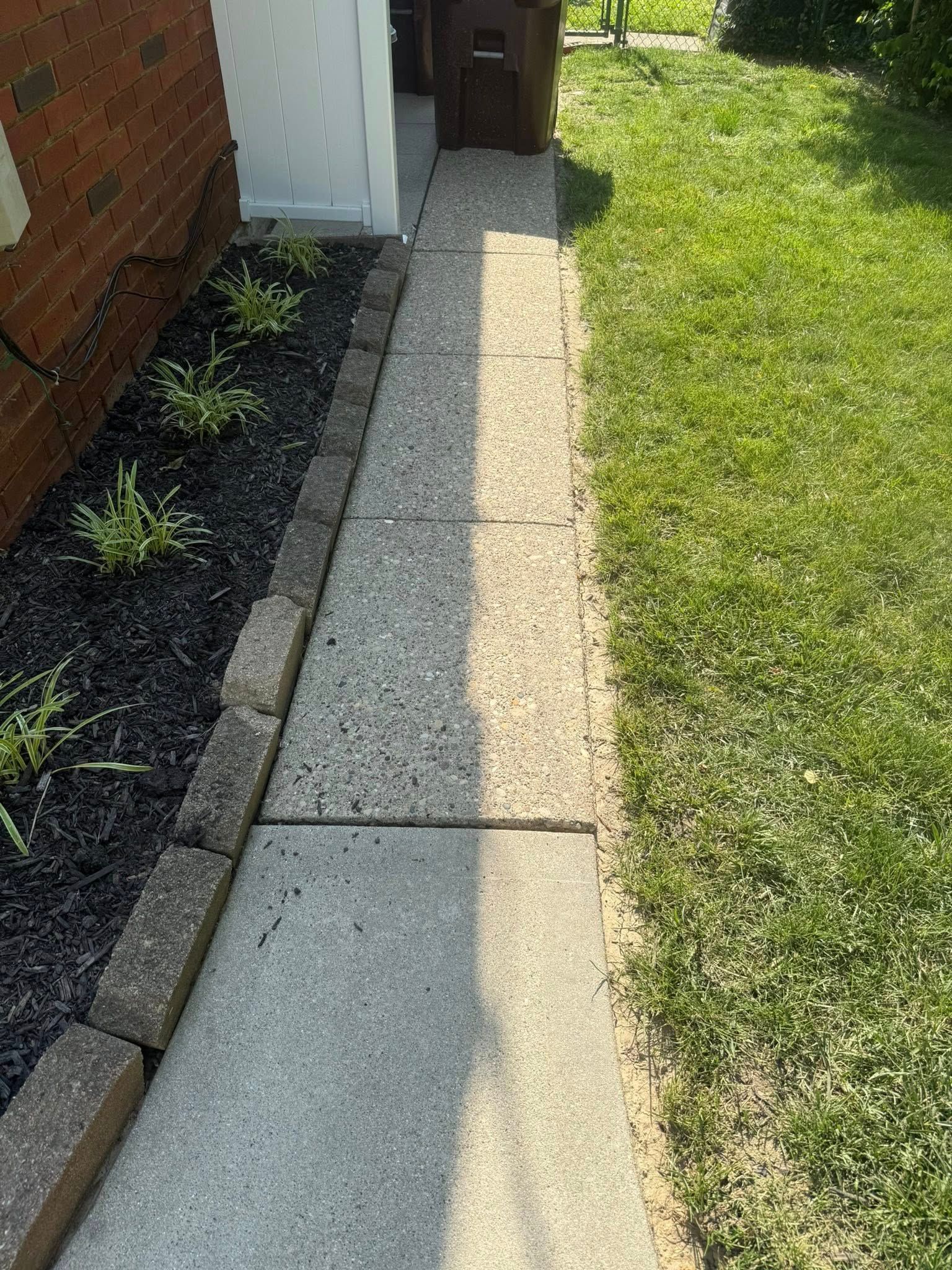 Narrow concrete walkway beside a brick house, with mulch border and green lawn in sunlight.