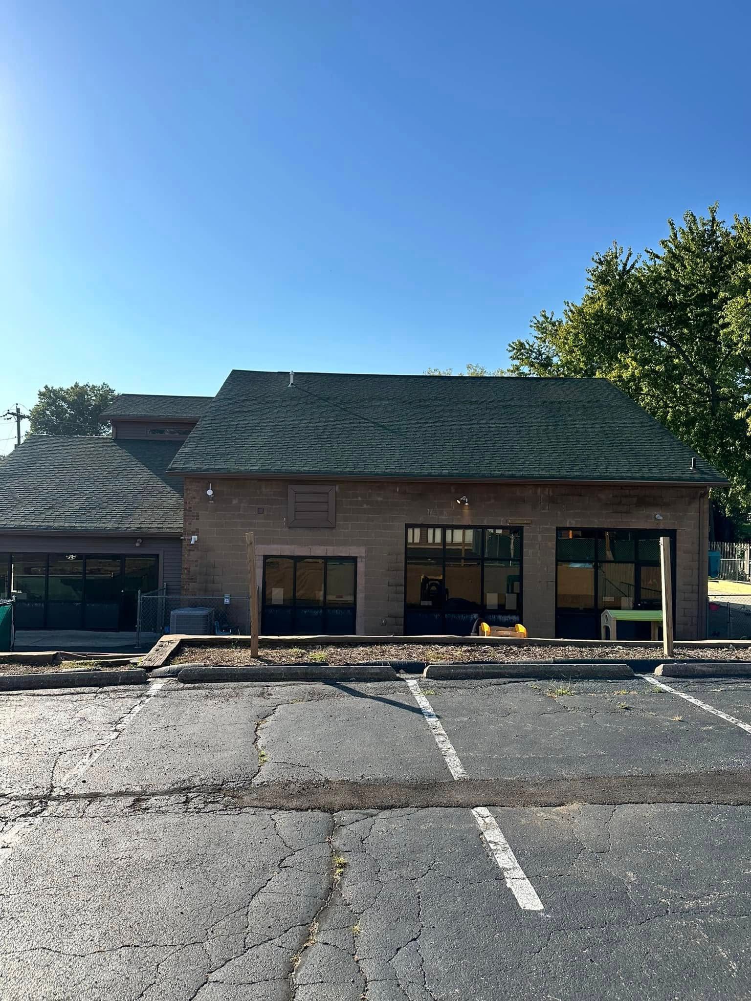Single-story brick building with a dark roof and large windows beside an empty parking lot under a blue sky