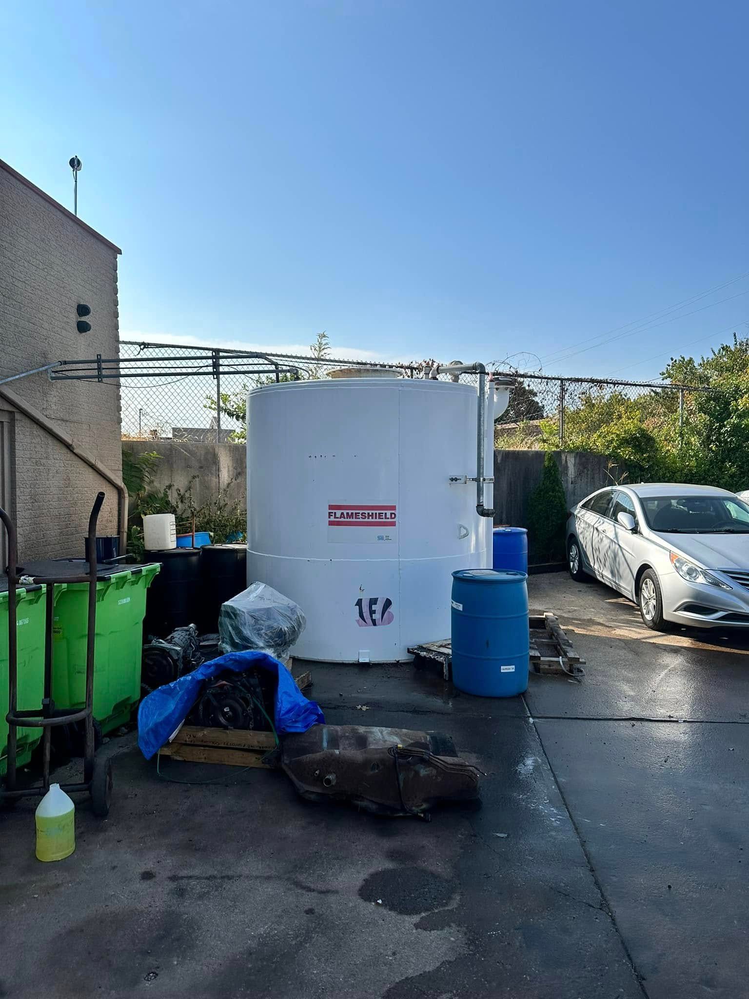 Outdoor work area with white tank, blue barrel, green bins, and a person crouching near a large rock.