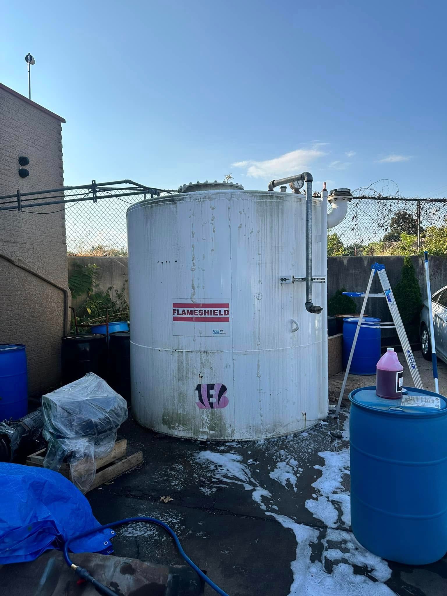 White cylindrical tank with warning label in a fenced industrial yard, surrounded by blue barrels and debris.