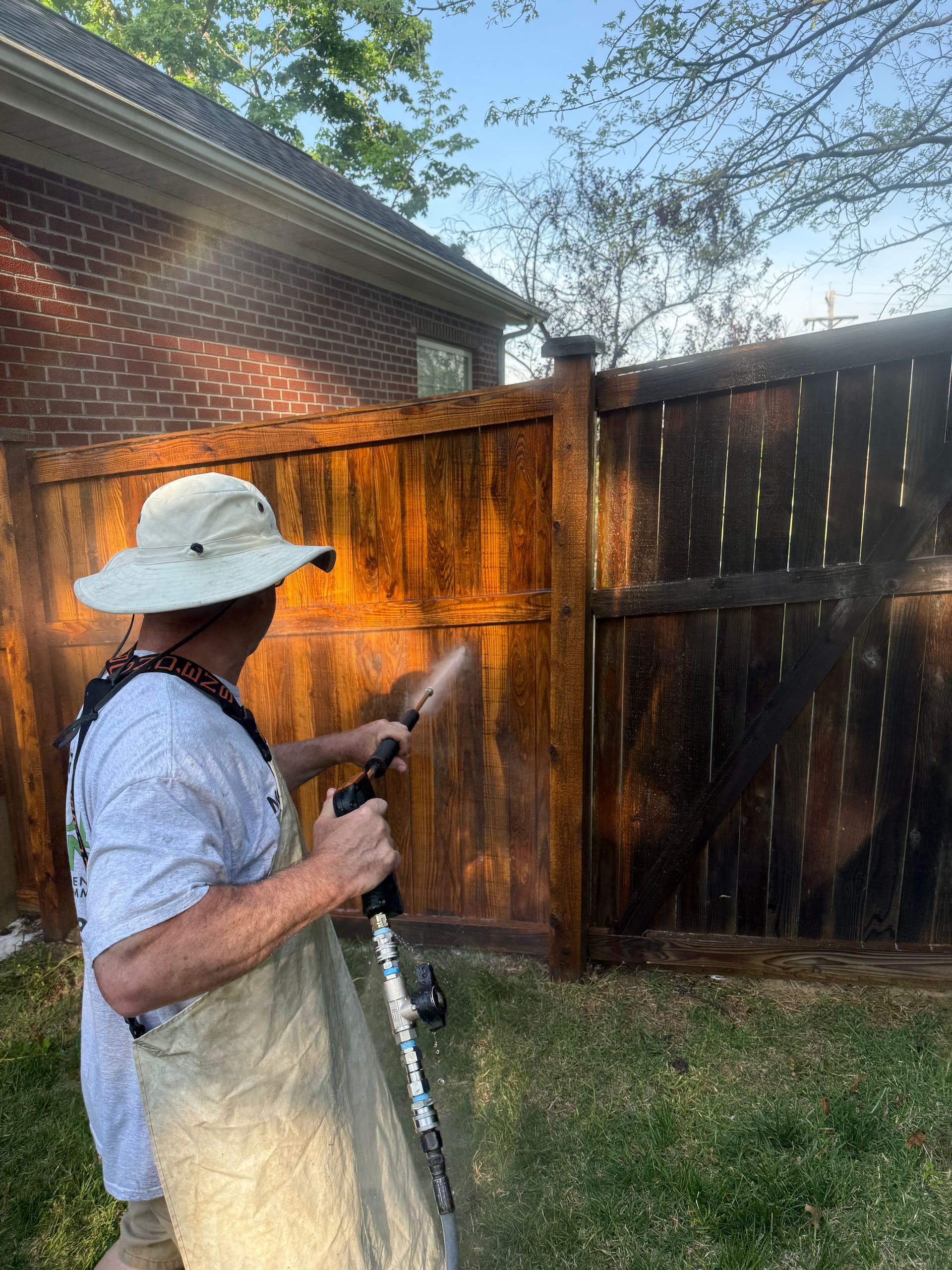 Person spraying a wooden fence with stain in a backyard, wearing a white hat and apron