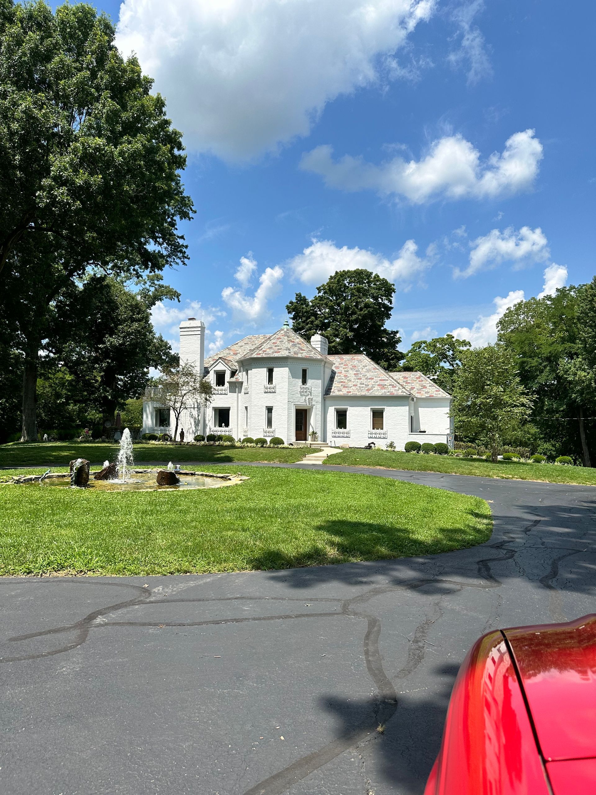 White country house beside a circular driveway, framed by trees and blue sky, with a red car in the foreground