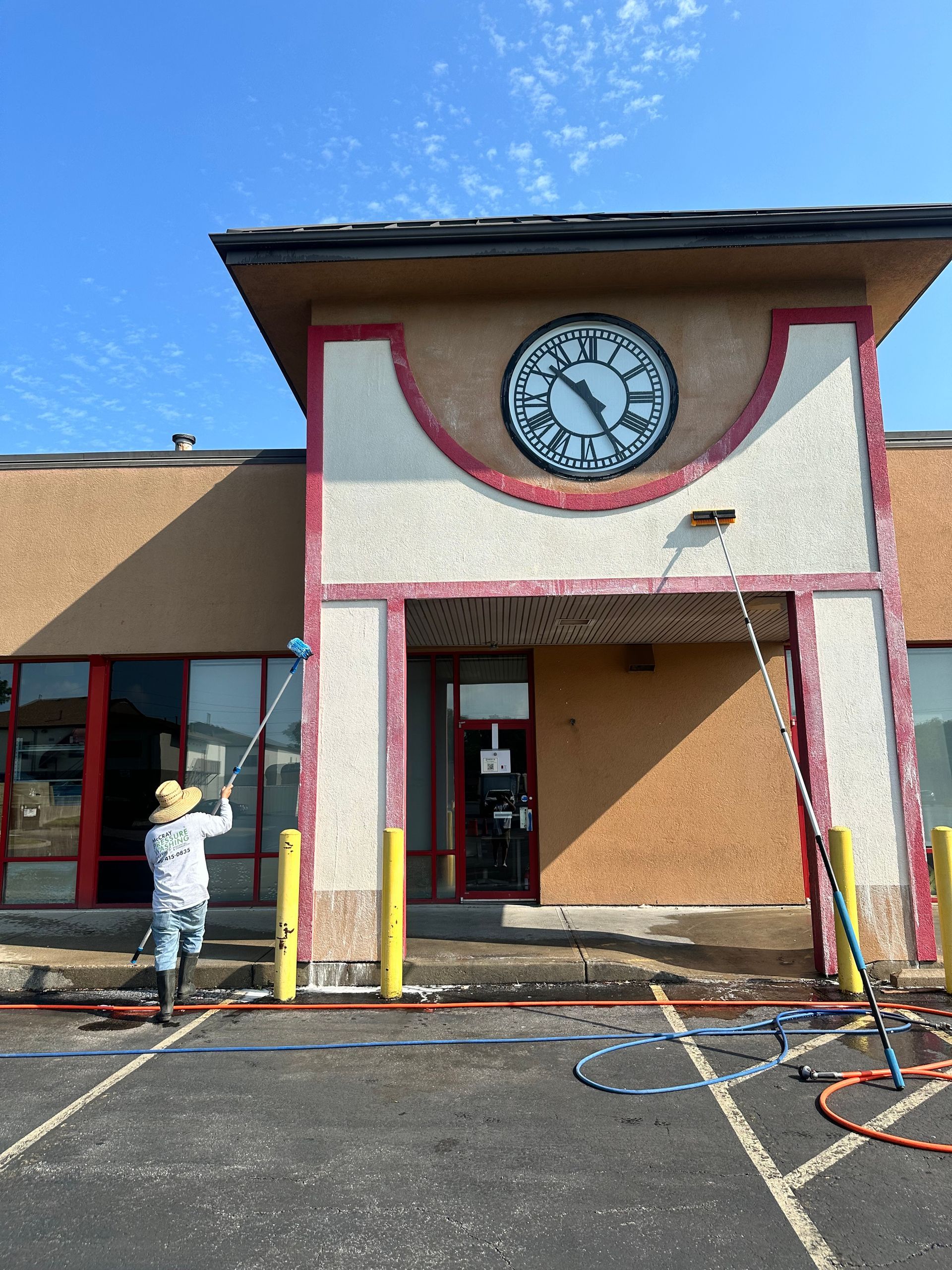 Person washing a storefront with a clock above the entrance and hoses on the ground